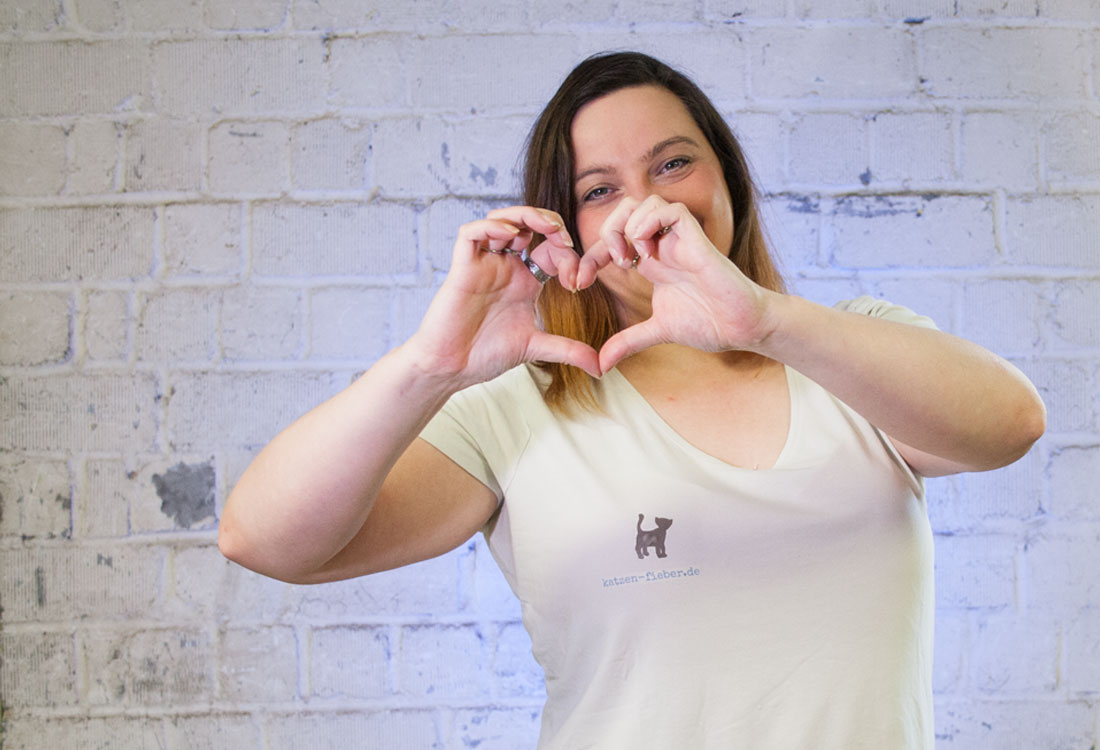 Eine brünette Frau im weißen T-Shirt mit Logo und Aufschrift katzen-fieber.de formt mit den Fingern ein Herz und lächelt in die Kamera